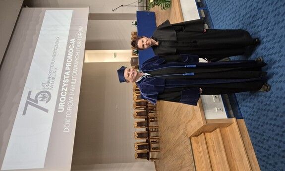 Two people in academic regalia stand on a stage during an honorary doctoral promotion ceremony.