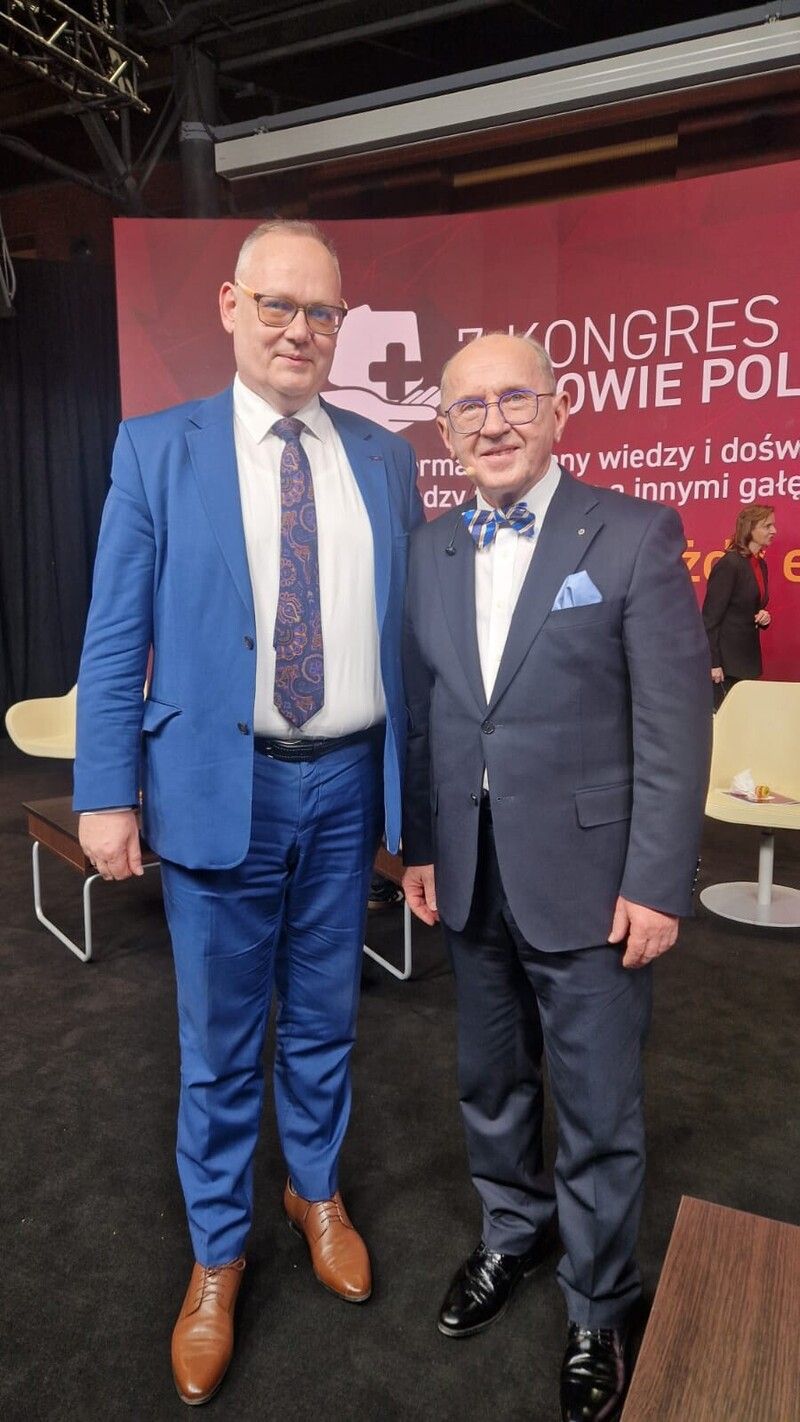 Two smiling men in suits stand together at a conference, looking at the camera.